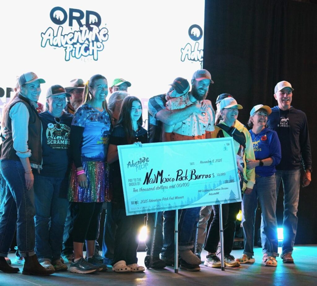 Group of people on a stage posing with a large ceremonial check at an Adventure Pitch event smiling and congratulating the winners